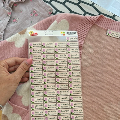 Mão a segurar uma folha de etiquetas verdes com o nome Laurelle sobre uma camisola cor-de-rosa